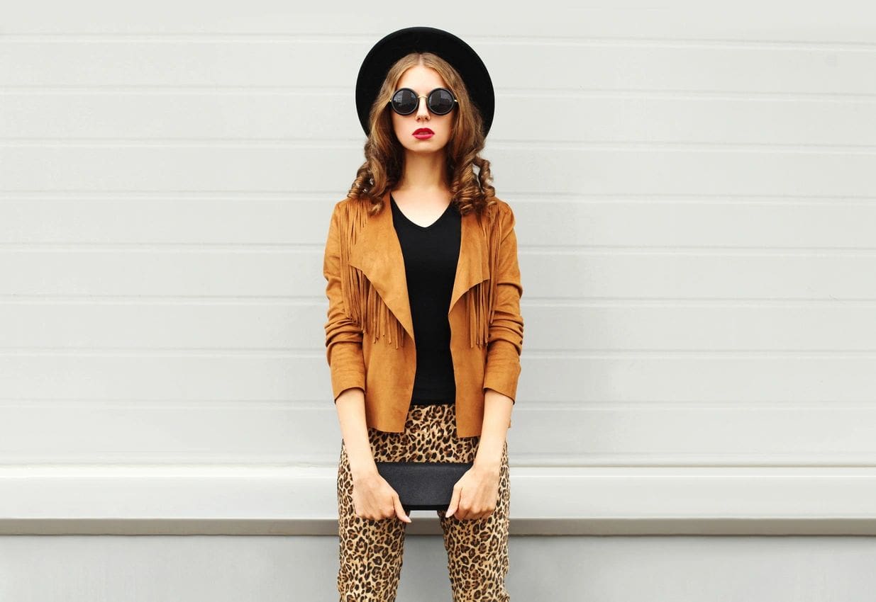 A woman in brown jacket and black hat holding purse.